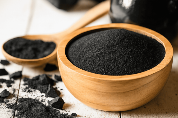 carbon black Natural carbon black powder in a wooden bowl and spoon, with scattered black carbon pieces on a white wooden surface – illustrating plant-based, clean-label natural black food color
